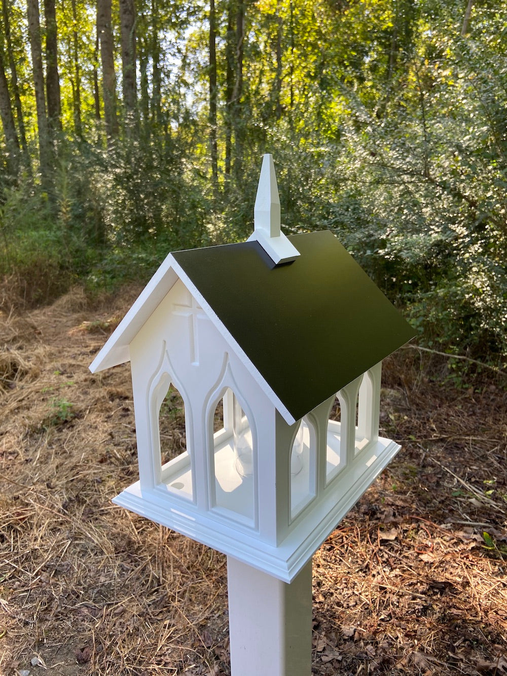 Black chapel birdfeeder mounted on white post with woods in the background