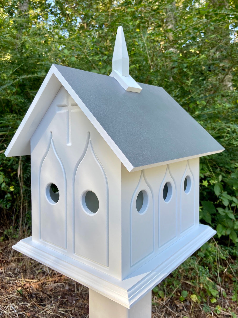 Gray chapel birdhouse on white post with woods in the background