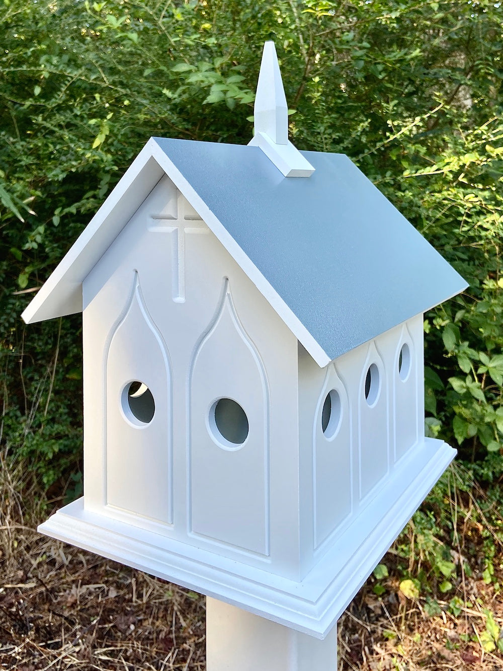 Light blue chapel birdhouse on white post with woods in the background