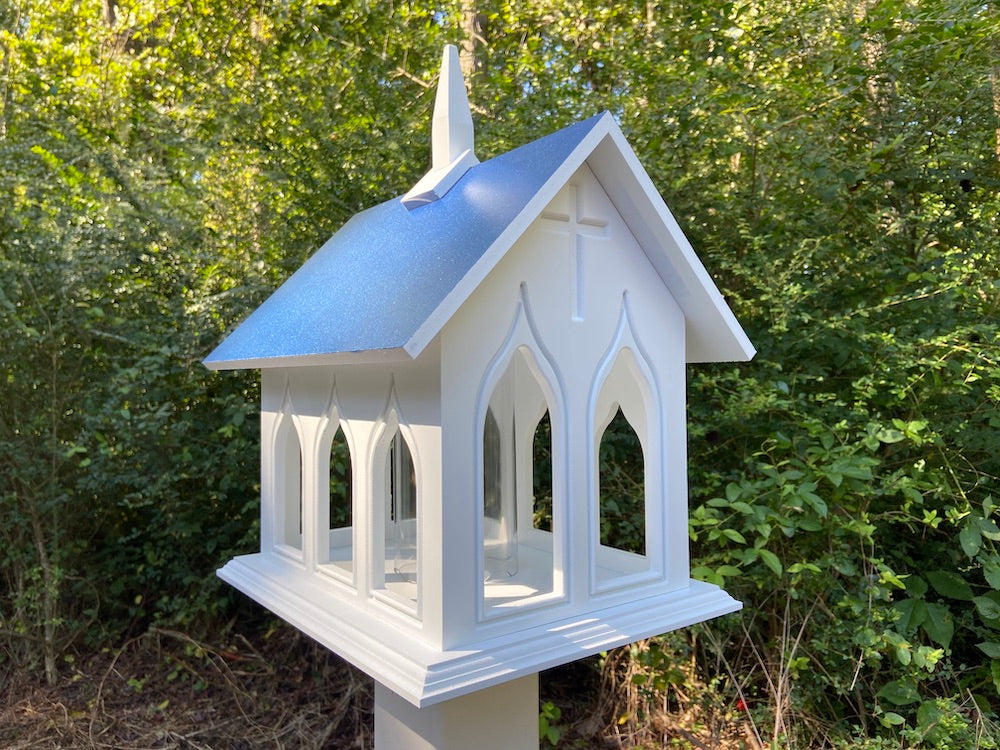 Cobalt blue chapel bird feeder mounted on white post with woods in the background