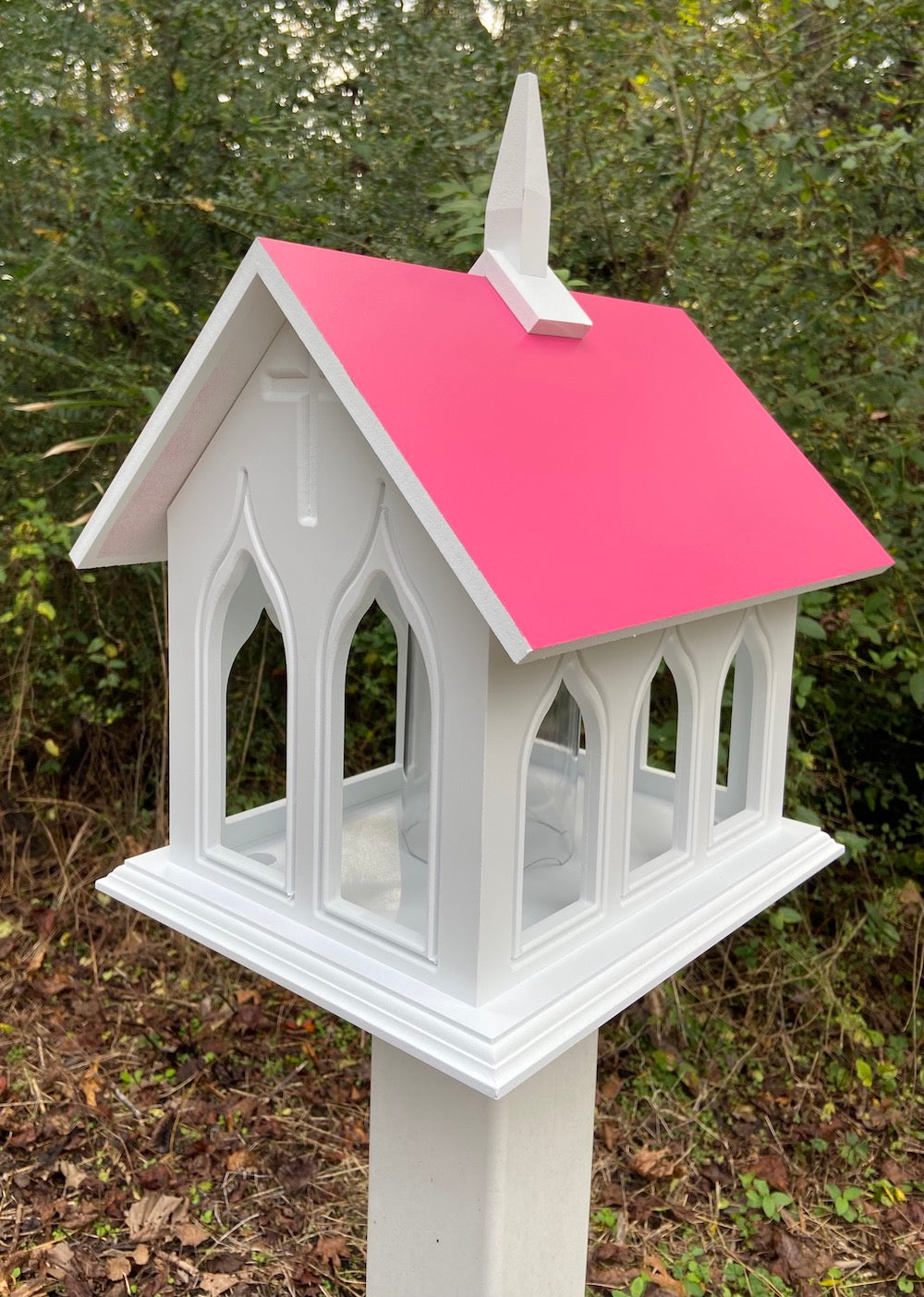 Pink chapel bird feeder mounted on white post with woods in the background