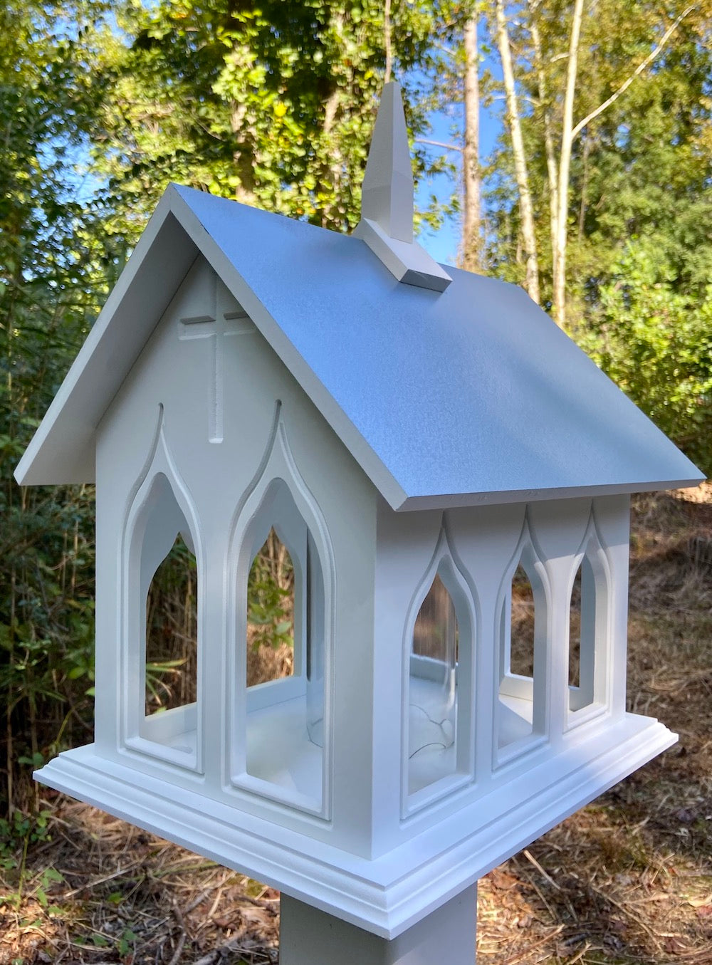 Light blue chapel bird feeder mounted on a white post with the woods in the background