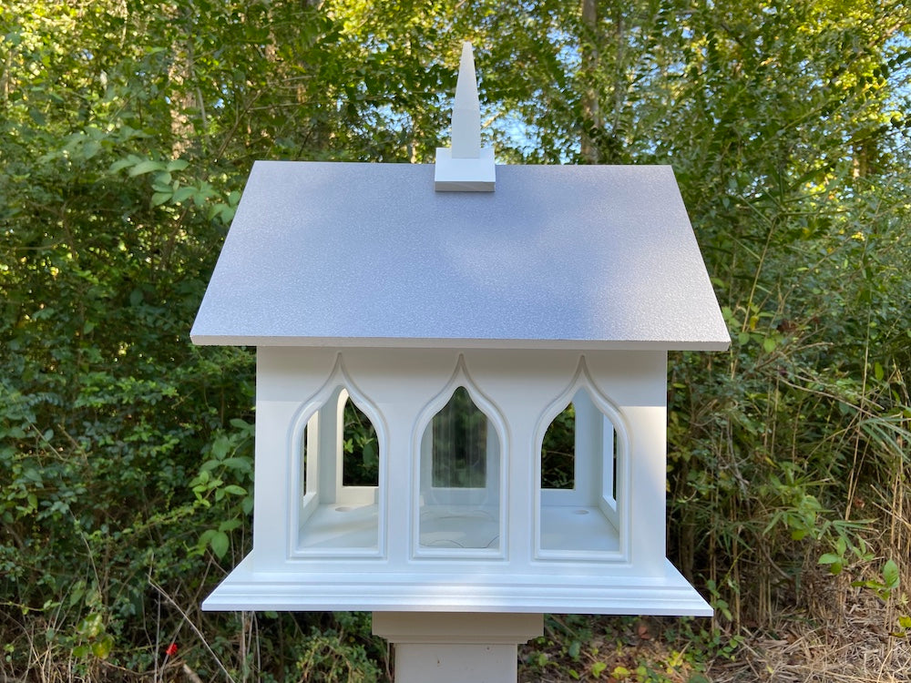 Gray chapel bird feeder mounted on a white post in the woods in the background